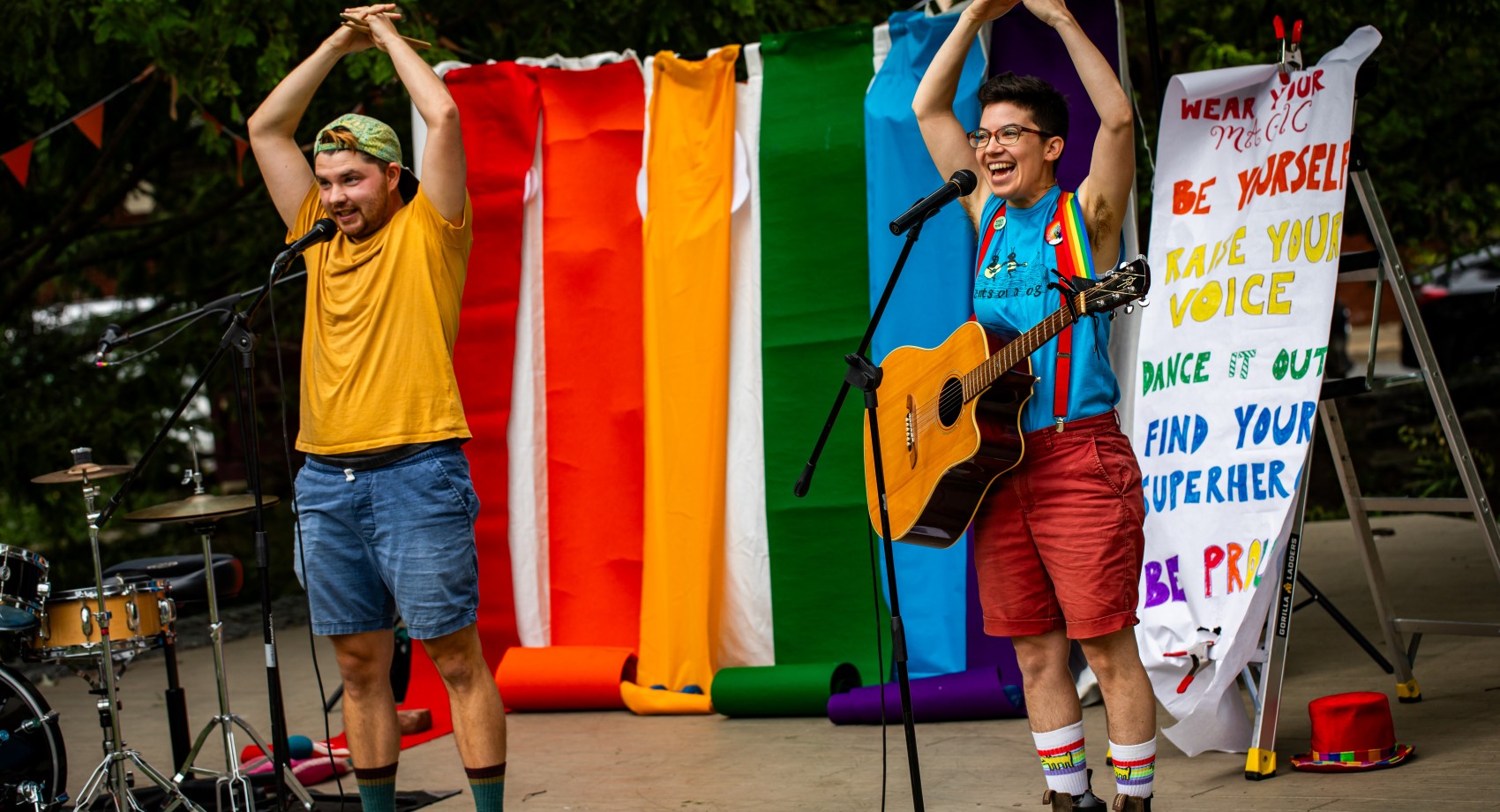 Two musicians raise their arms above their heads in celebration. One holds a guitar, one stands next to a drumset.