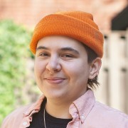 Nonbinary white person wearing an orange hat smiles into the camera