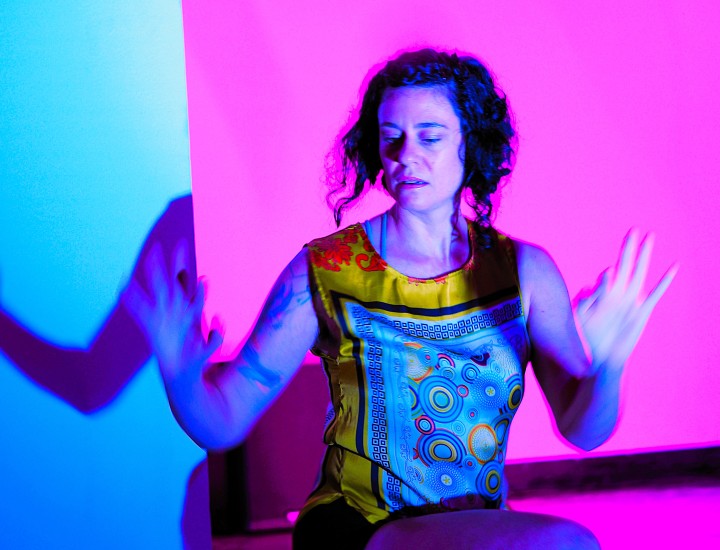 Nicole, a white Jewish woman with bob-length, assymetrical curly hair, is dancing in a pink glowing light. Her torso is slightly twisted with her hands up and gaze down. She is wearing an iridescent yellow tank-dress. Her arms are casting shadows against a white gallery wall.