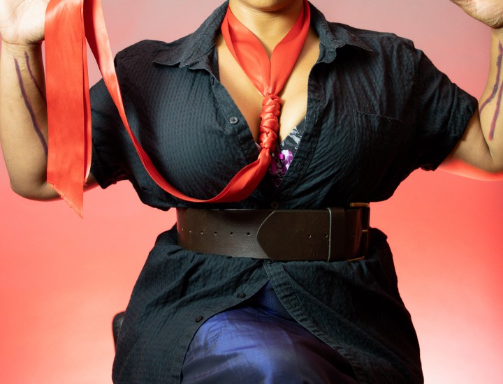 Photo of a light skinned black femme in a black button up shirt and dark blue long skirt holding their hands up with a soft red noose as a necklace
