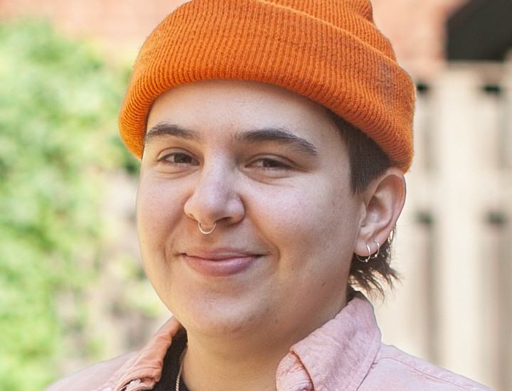 Nonbinary white person wearing an orange hat smiles into the camera