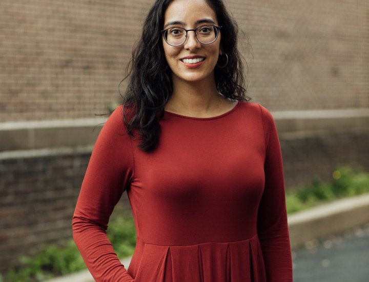 A Latinx cis woman with wavy hair and smiling, wearing purple glasses and a red dress